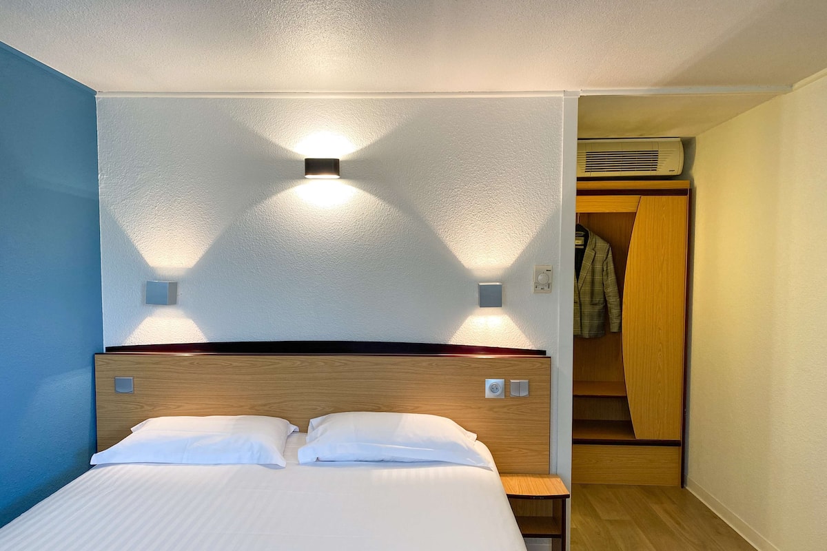 Habitación de hotel con cama, controles junto a la cama, una mesita pequeña y un armario con una prenda colgada.