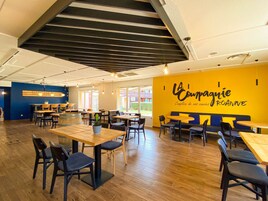 Un interior de cafetería moderno con mesas y sillas de madera, una pared de acento amarilla con un letrero y un techo inclinado con vigas a la vista.