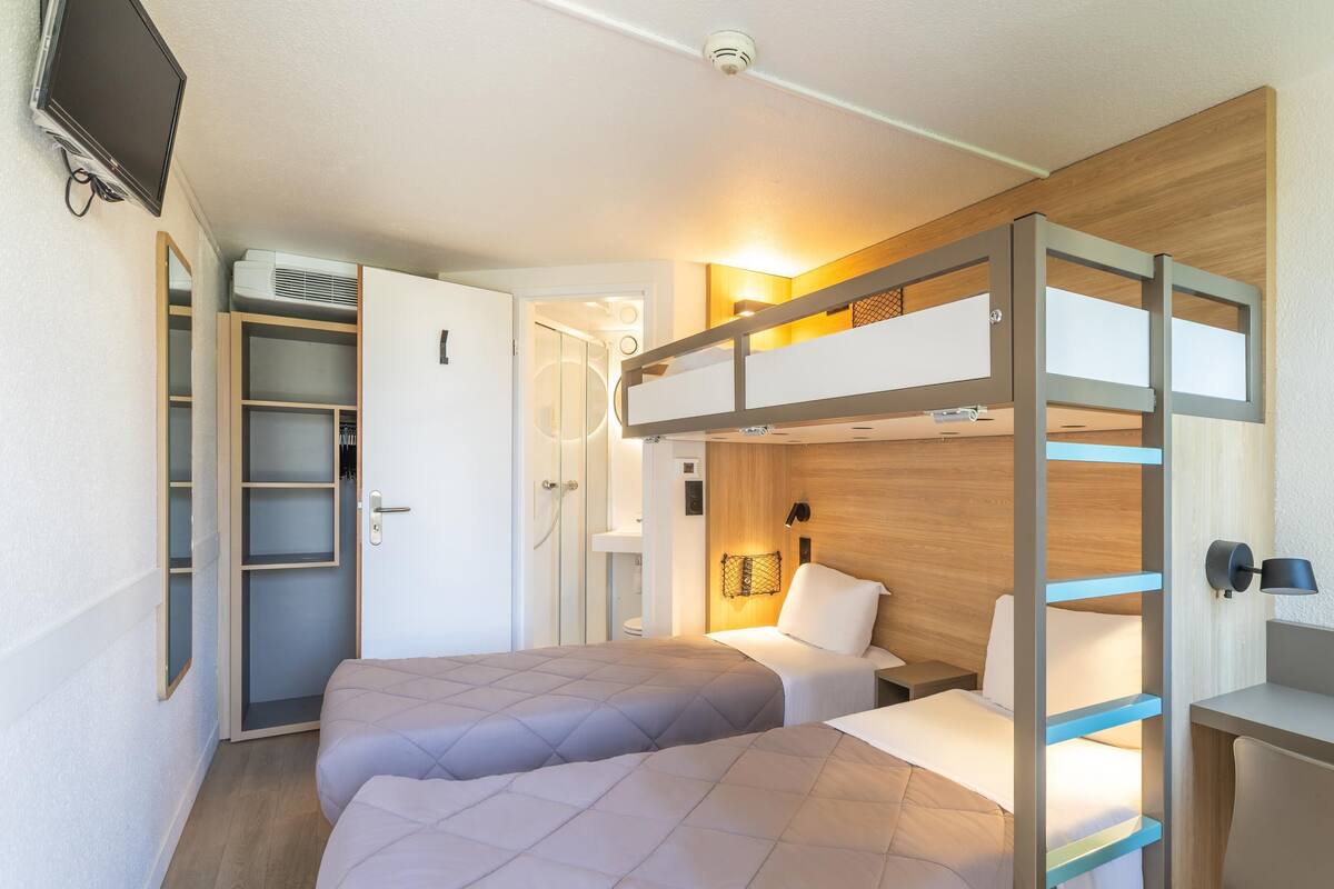 Een kleine hotelkamer met een stapelbed, een bureau, een kleine badkamer en een aan de muur bevestigde televisie.