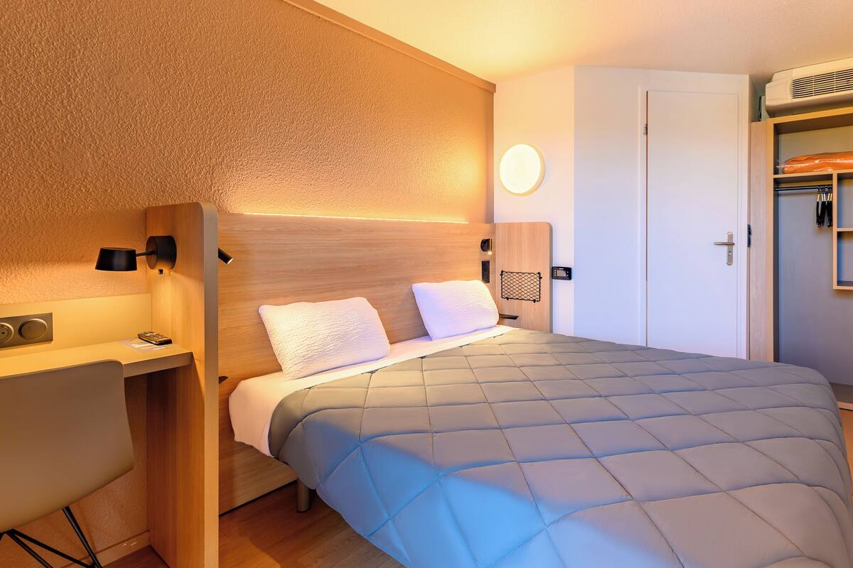 Een kleine hotelkamer met een bed, nachtkastje, lamp en een kledingkast met planken.