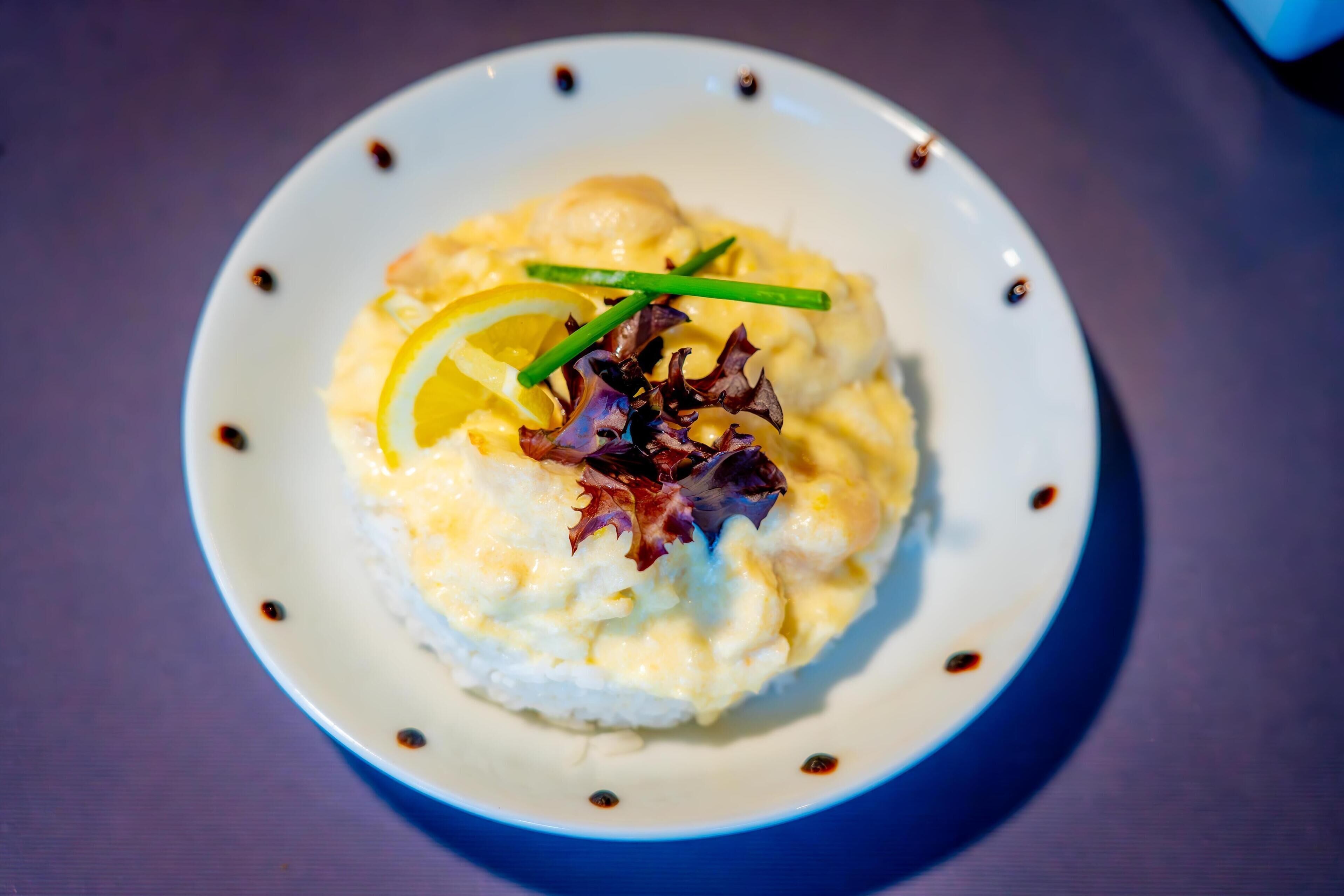 Un plato de comida con una base cremosa, coronado con una rodaja de limón y cebollín verde, y adornado con una hoja de lechuga morada.