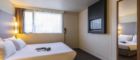 Habitación de hotel con cama, ventana con cortinas, televisor montado en la pared y un espejo.
