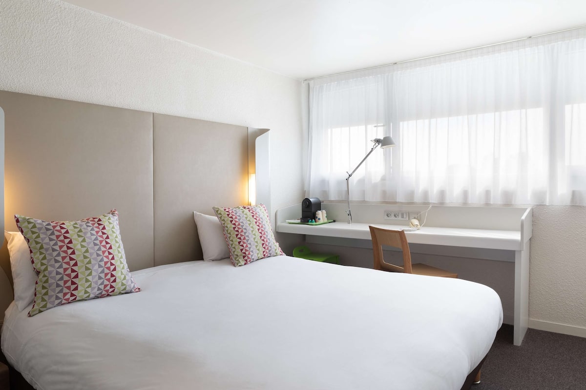 Une chambre d’hôtel avec un grand lit, un bureau avec une chaise, une fenêtre avec des rideaux légers et une lampe sur le bureau.