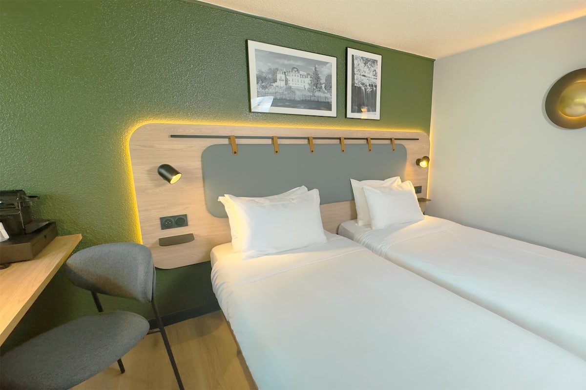 Ein Hotelzimmer mit zwei Einzelbetten, einem Schreibtisch mit Stuhl, einem kleinen Tisch und einer Wandlampe.