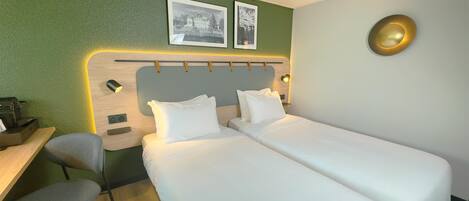 Habitación de hotel con dos camas individuales, un escritorio con silla, una mesita y una lámpara de pared.