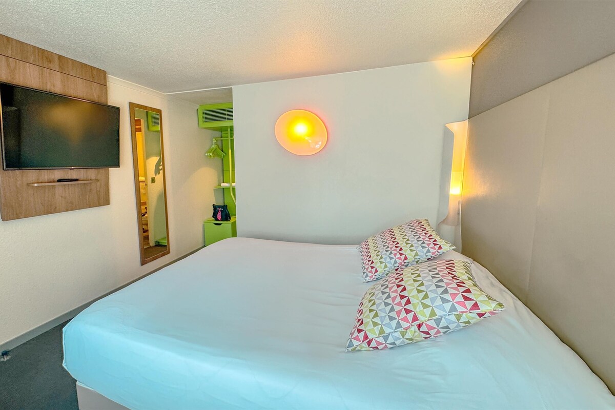 A hotel room with a bed, a TV mounted on the wall, a mirror, and a wall-mounted light fixture.