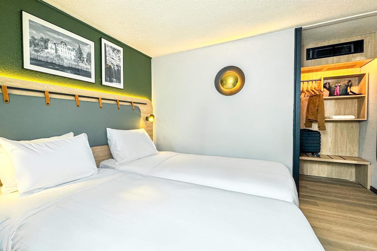 A hotel room with a large bed, a headboard with a light fixture, and a wardrobe with hanging clothes.