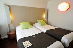 Two single beds with white bedding and green pillows, a nightstand with a lamp and power outlet, and a wall-mounted light fixture.