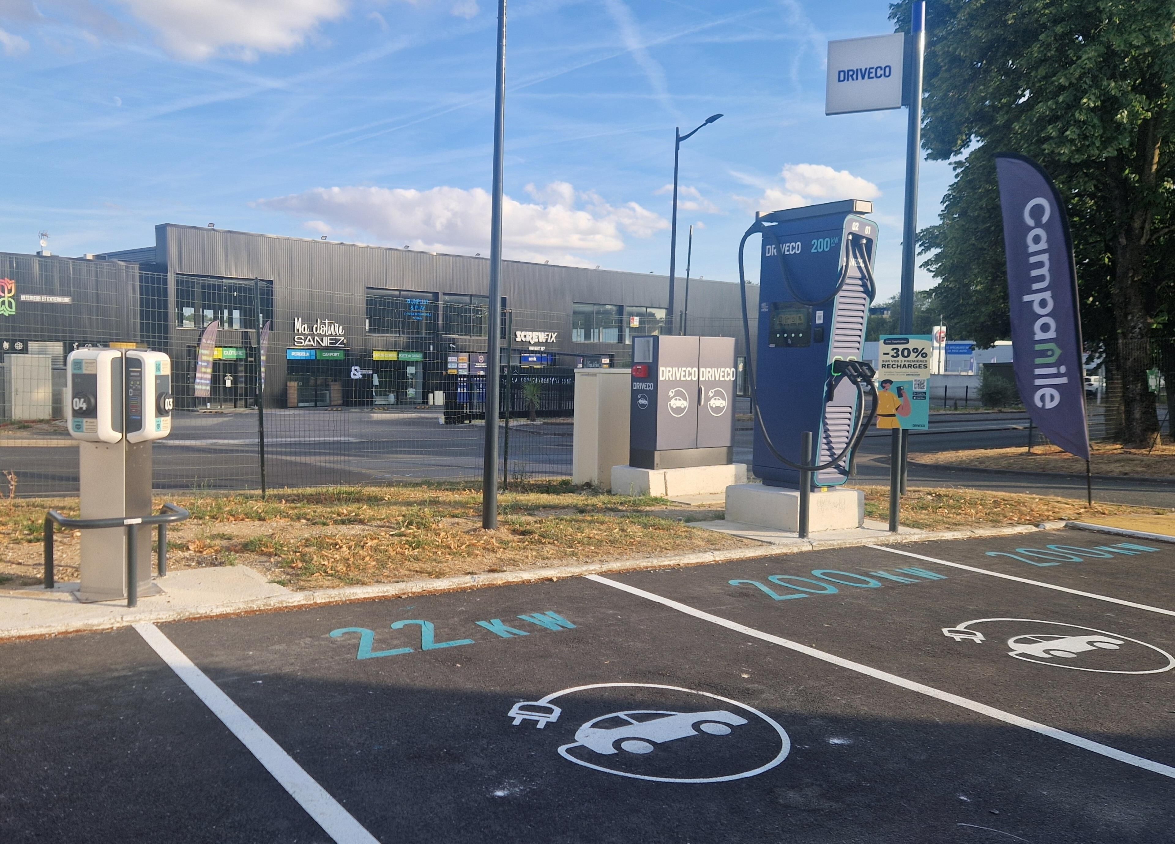 Un área de estacionamiento con estaciones de carga para vehículos eléctricos, un área de servicio con acceso para vehículos y un edificio con señalización.