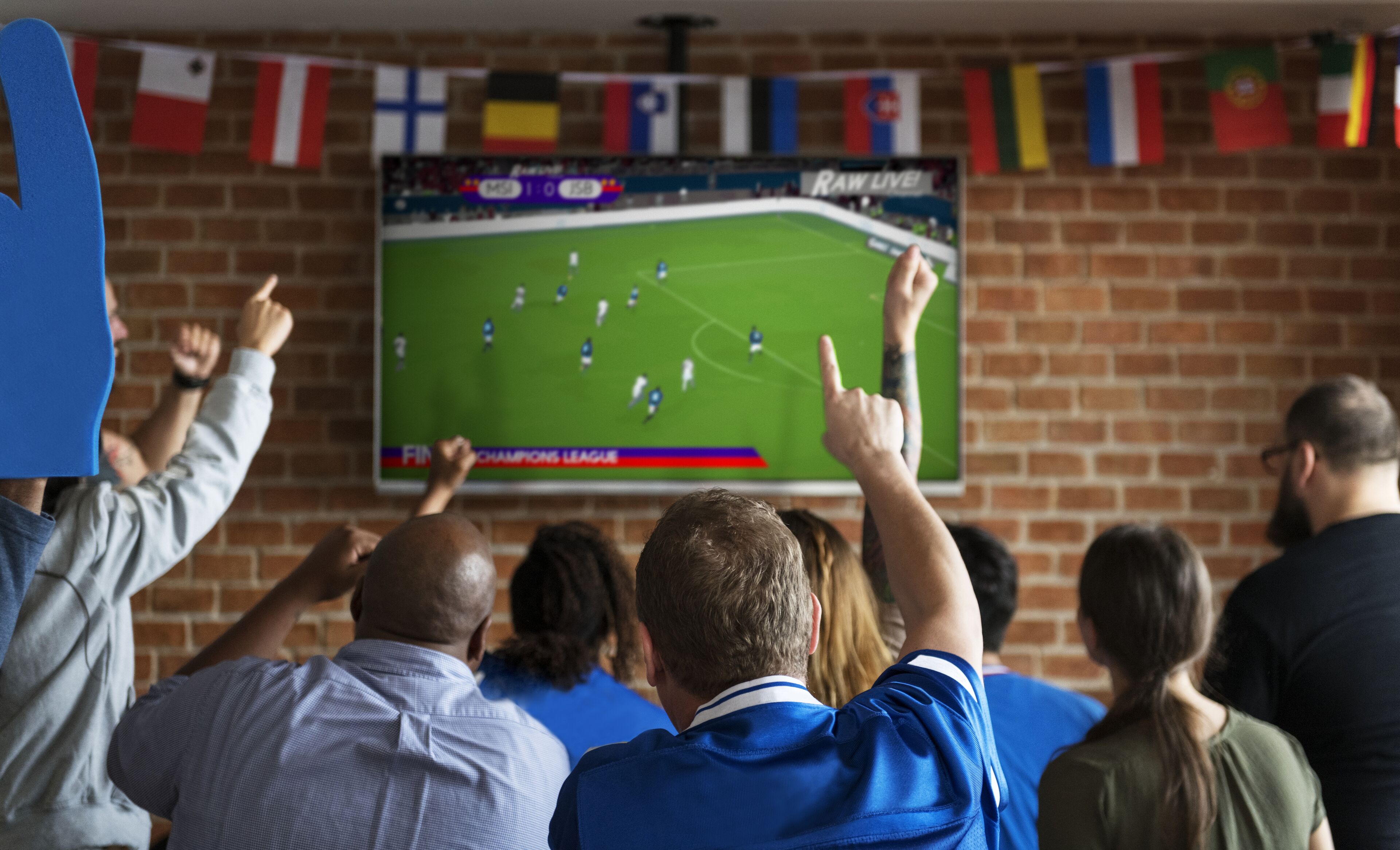 Een groep mensen kijkt naar een sportwedstrijd op een scherm, met een bakstenen muur en vlaggen op de achtergrond.