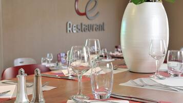 A dining table set with wine glasses, water glasses, and cutlery, with a large vase and a sign reading "Le Restaurant" in the background.