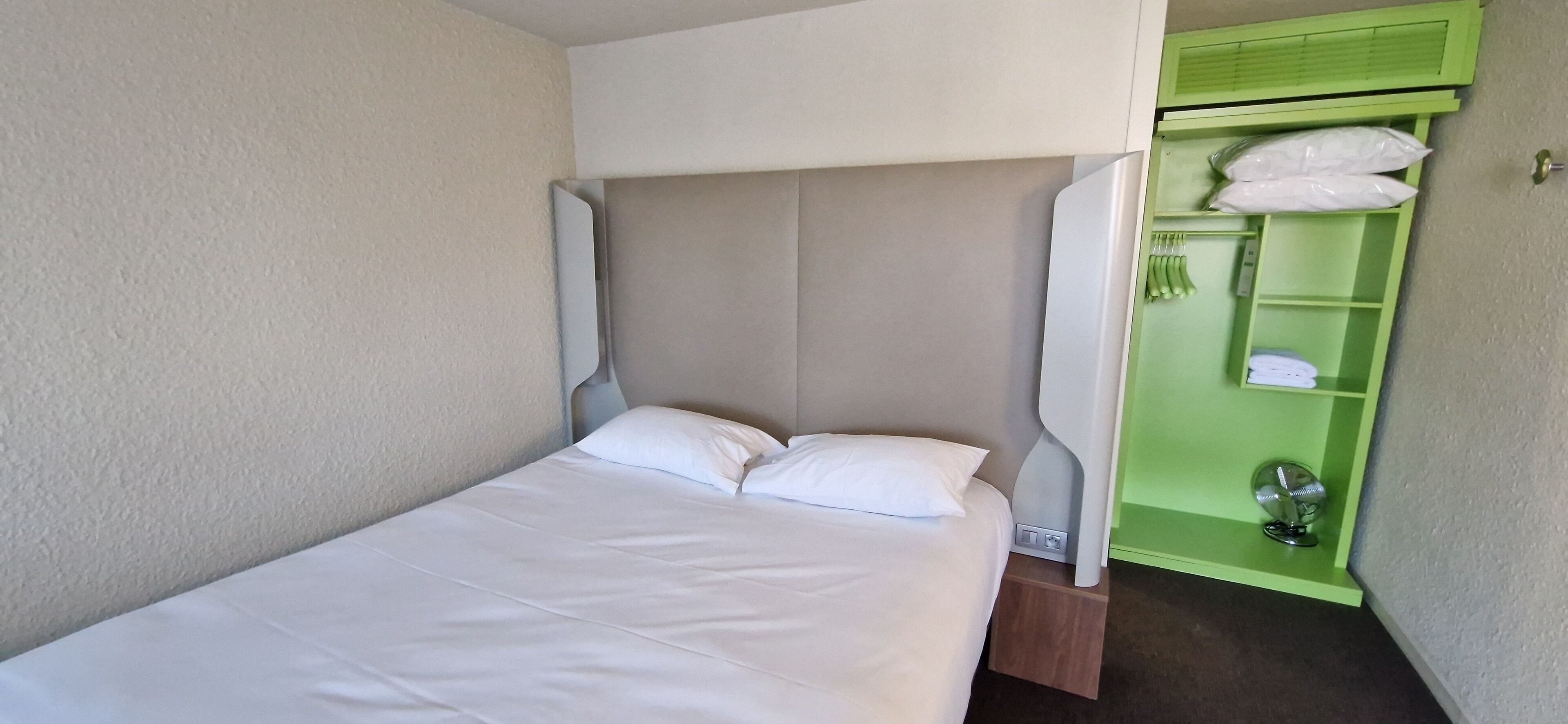 Un lit simple avec une literie blanche, une tête de lit et une table de chevet. Une étagère verte avec un ventilateur est visible sur la droite.