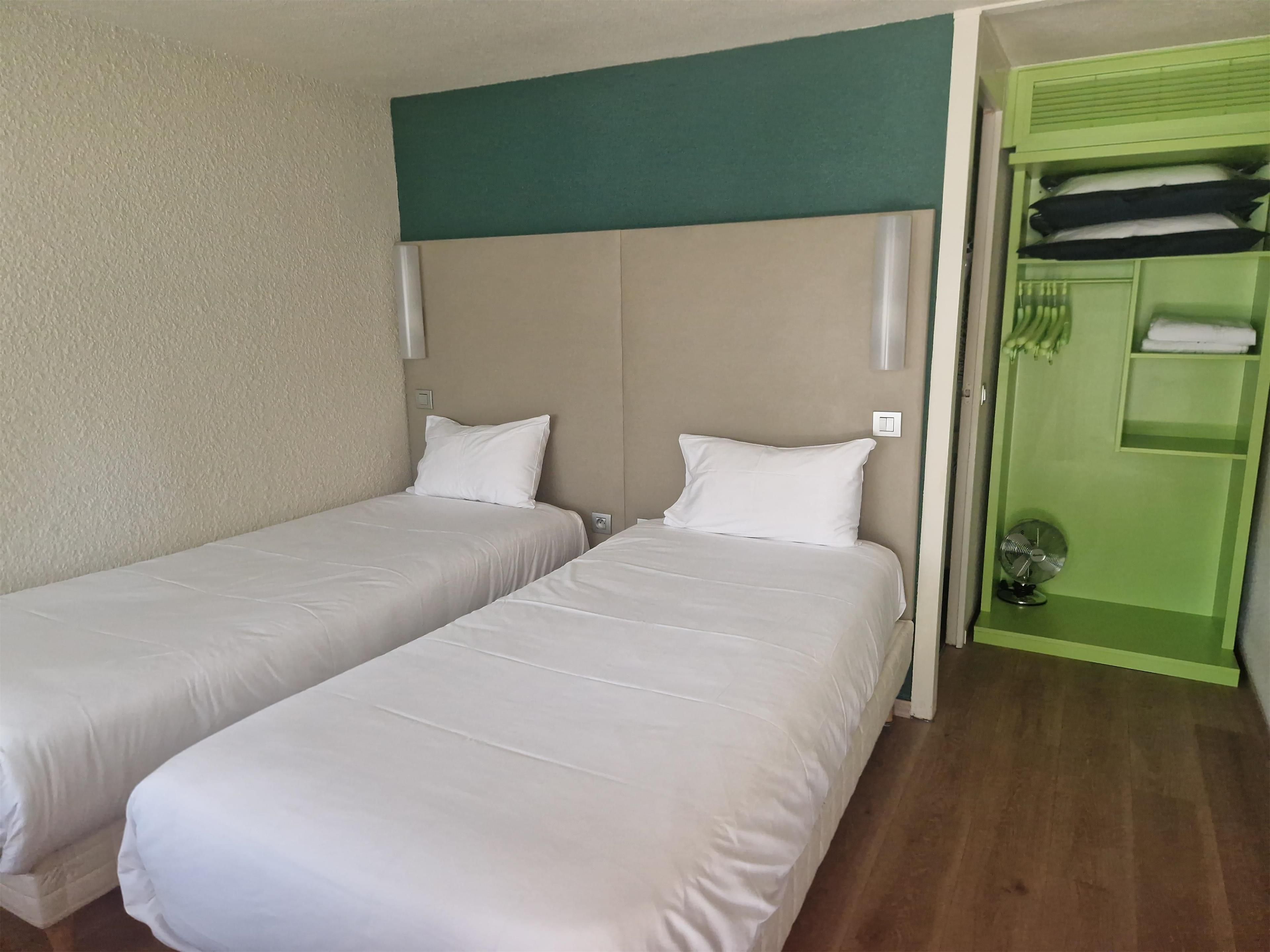 A hotel room with two single beds, a green wardrobe, and a small fan.