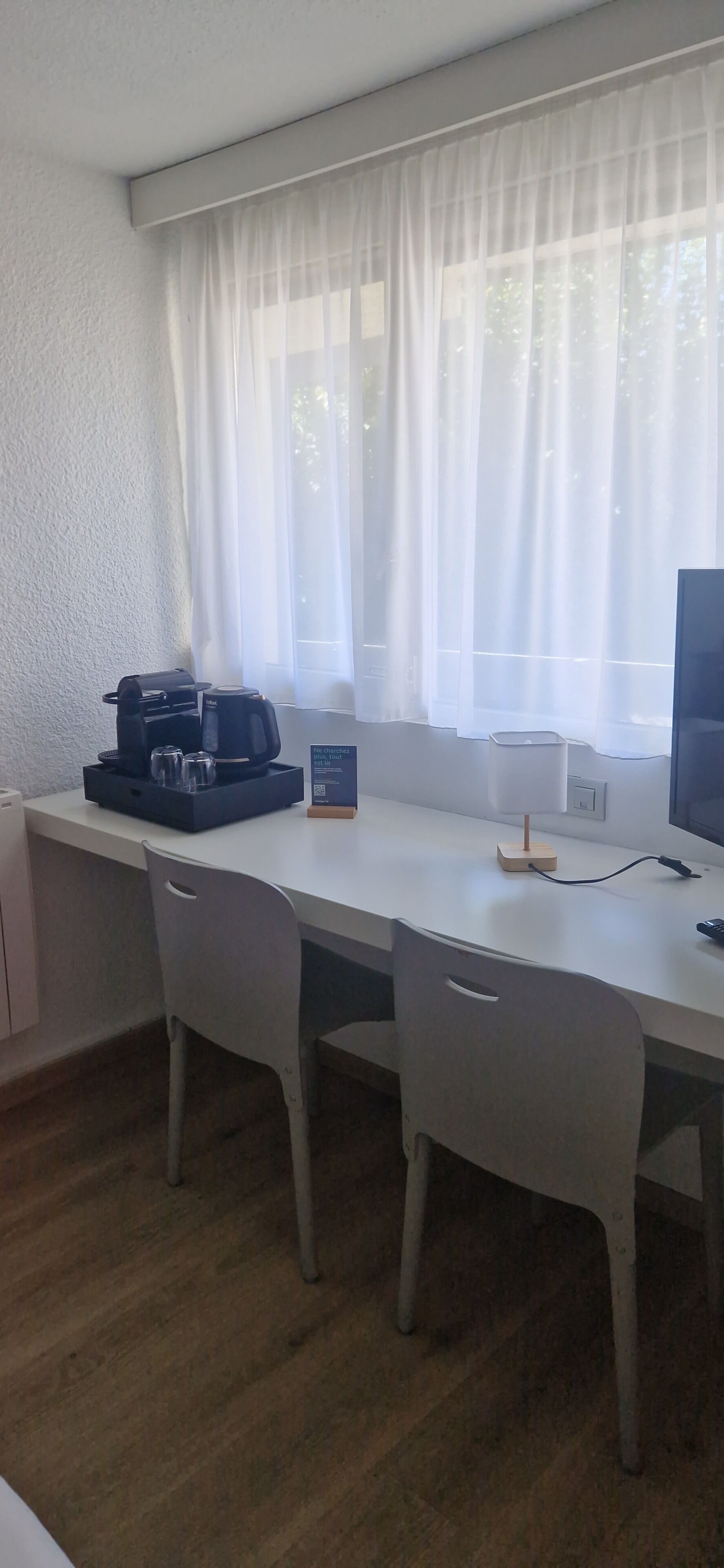 A small, clean room with a white desk, two chairs, a coffee maker, a lamp, and a TV.