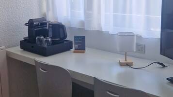 A small, clean room with a white desk, two chairs, a coffee maker, a lamp, and a TV.