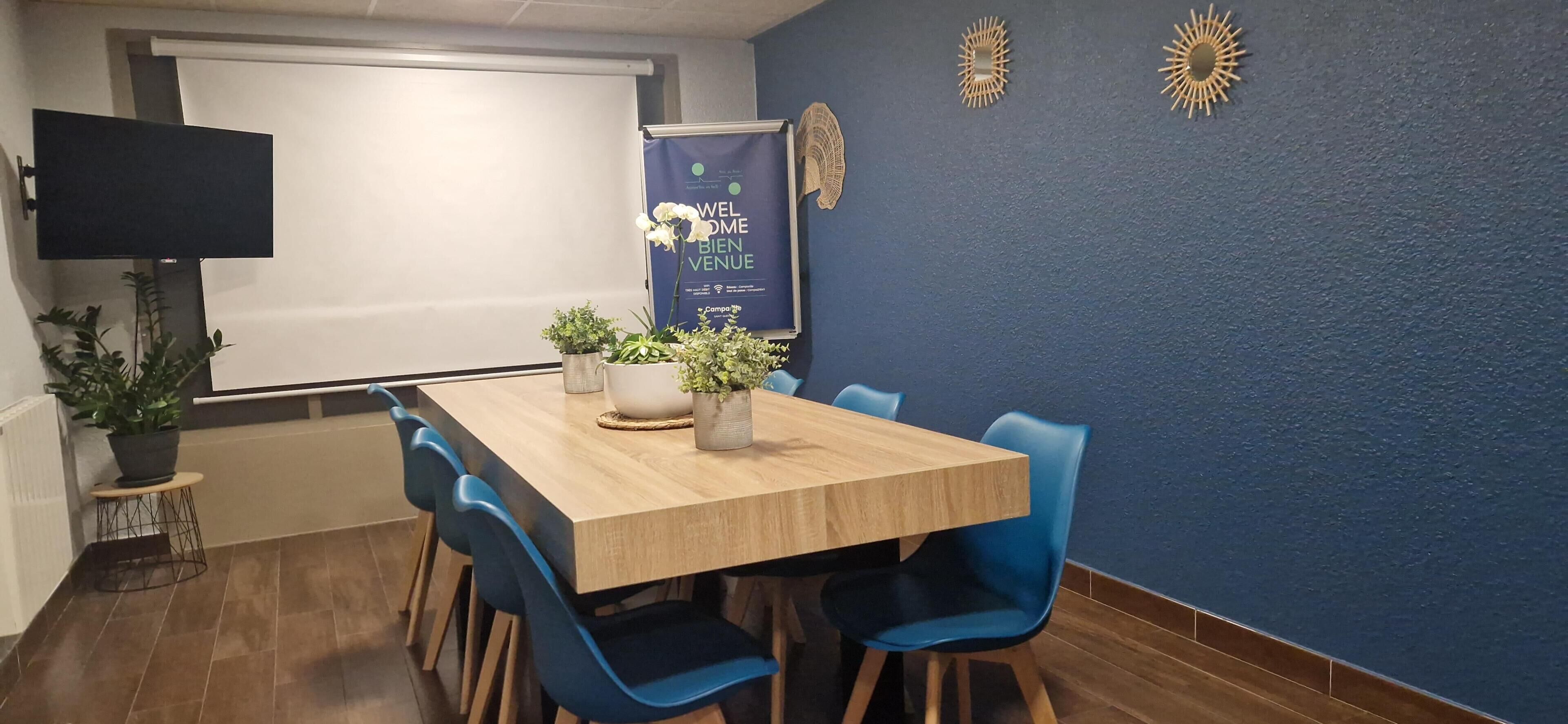 Une salle de conférence avec une table en bois, des chaises bleues, des plantes en pot et une banderole sur laquelle on peut lire : « BIENVENUE ».