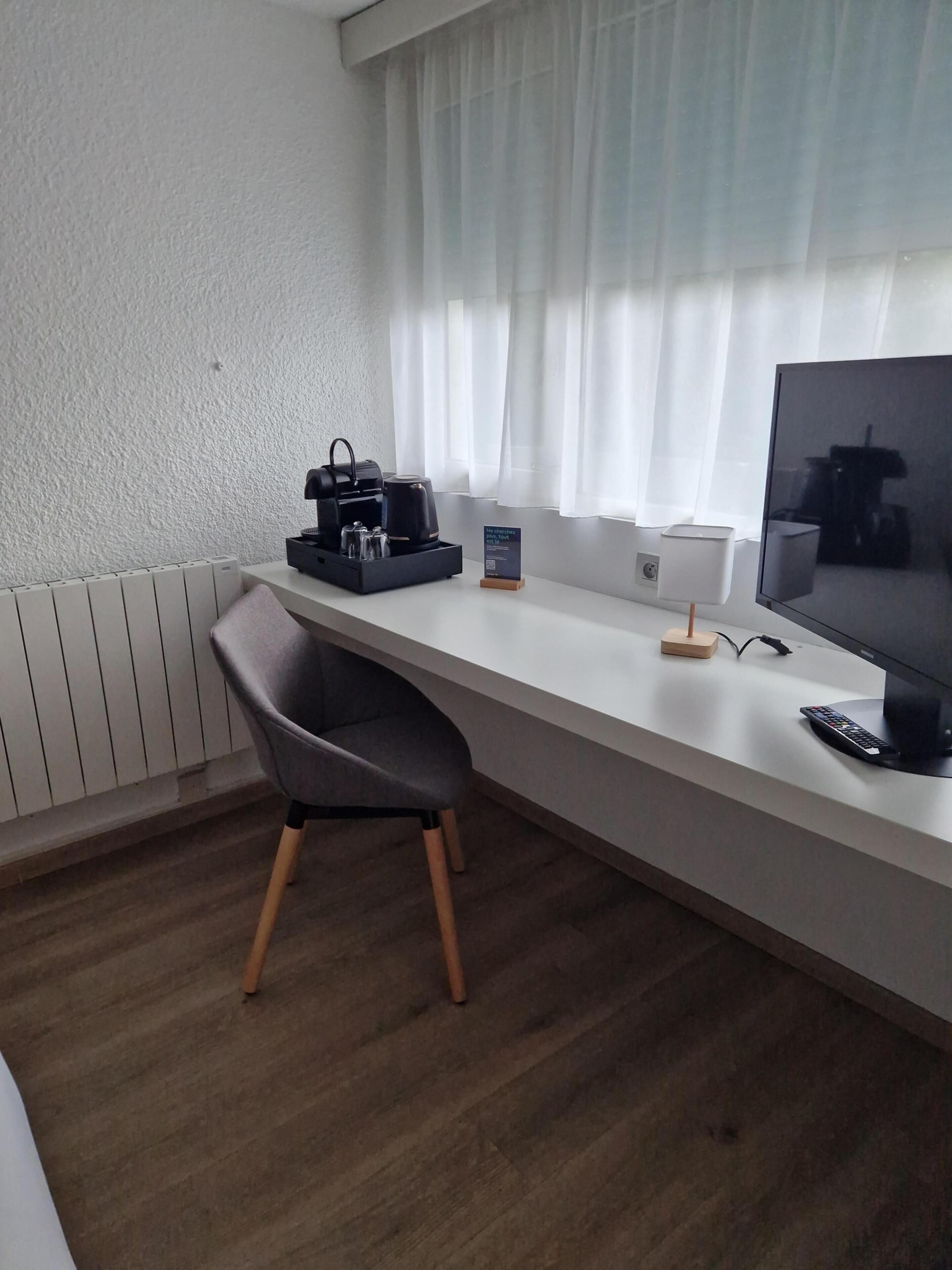 Una habitación de hotel moderna con un escritorio blanco, una silla negra, un televisor de pantalla plana, una cafetera y un pequeño cartel.