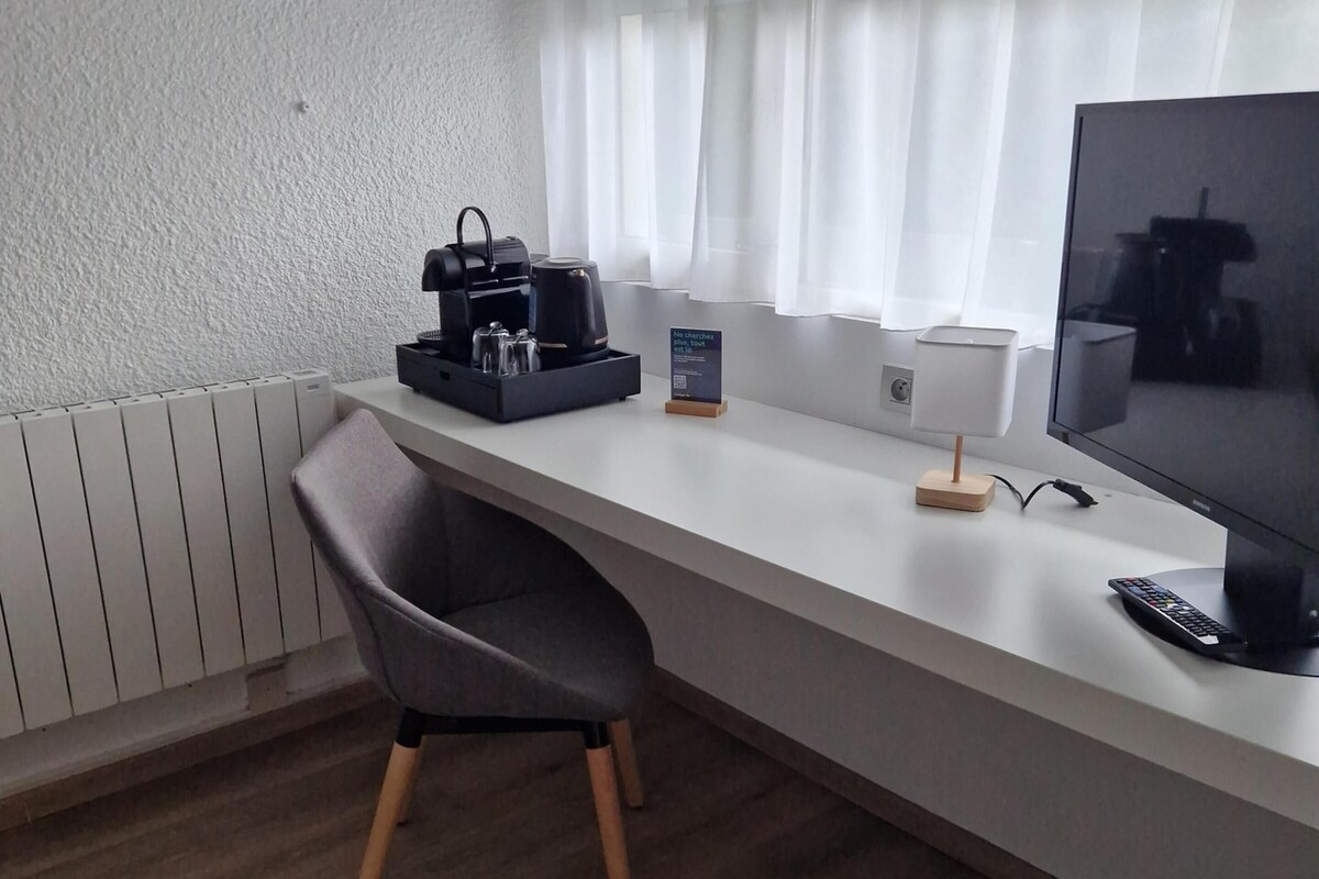 Una habitación de hotel moderna con un escritorio blanco, una silla negra, un televisor de pantalla plana, una cafetera y un pequeño cartel.