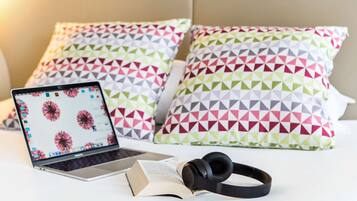 Una laptop sobre la cama con un libro y auriculares, rodeada de almohadones geométricos de colores.