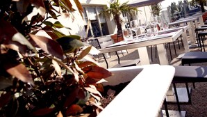 An outdoor dining area with tables and chairs, a palm tree, and a building with a sign in the background.