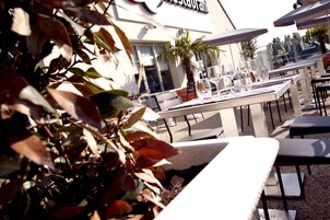 Un espace extérieur aménagé pour les repas, avec des tables et des chaises, un palmier et un bâtiment dont on aperçoit une enseigne au fond.