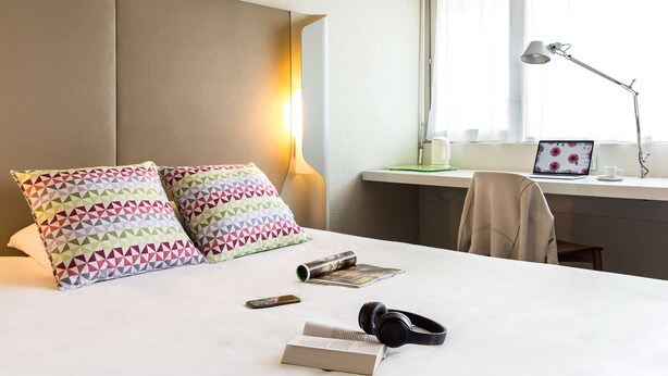 A neatly made bed with patterned pillows, a bedside table with a lamp, a laptop, and a chair.