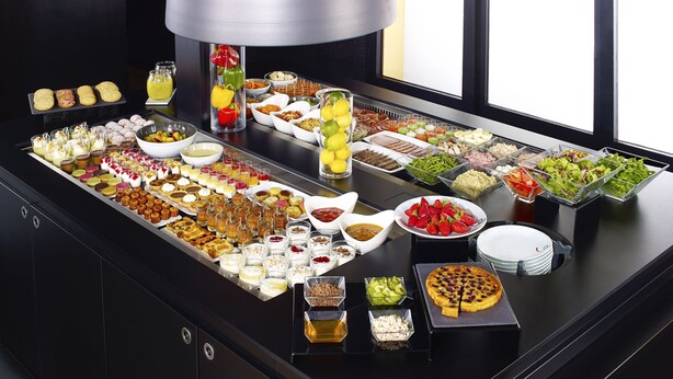 A buffet with a variety of food items and beverages, including fruits, salads, and desserts, arranged on a black table with a large lamp above. - KYRIAD Sedan (Sedan)