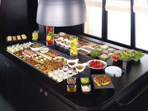 A buffet with a variety of food items and beverages, including fruits, salads, and desserts, arranged on a black table with a large lamp above.