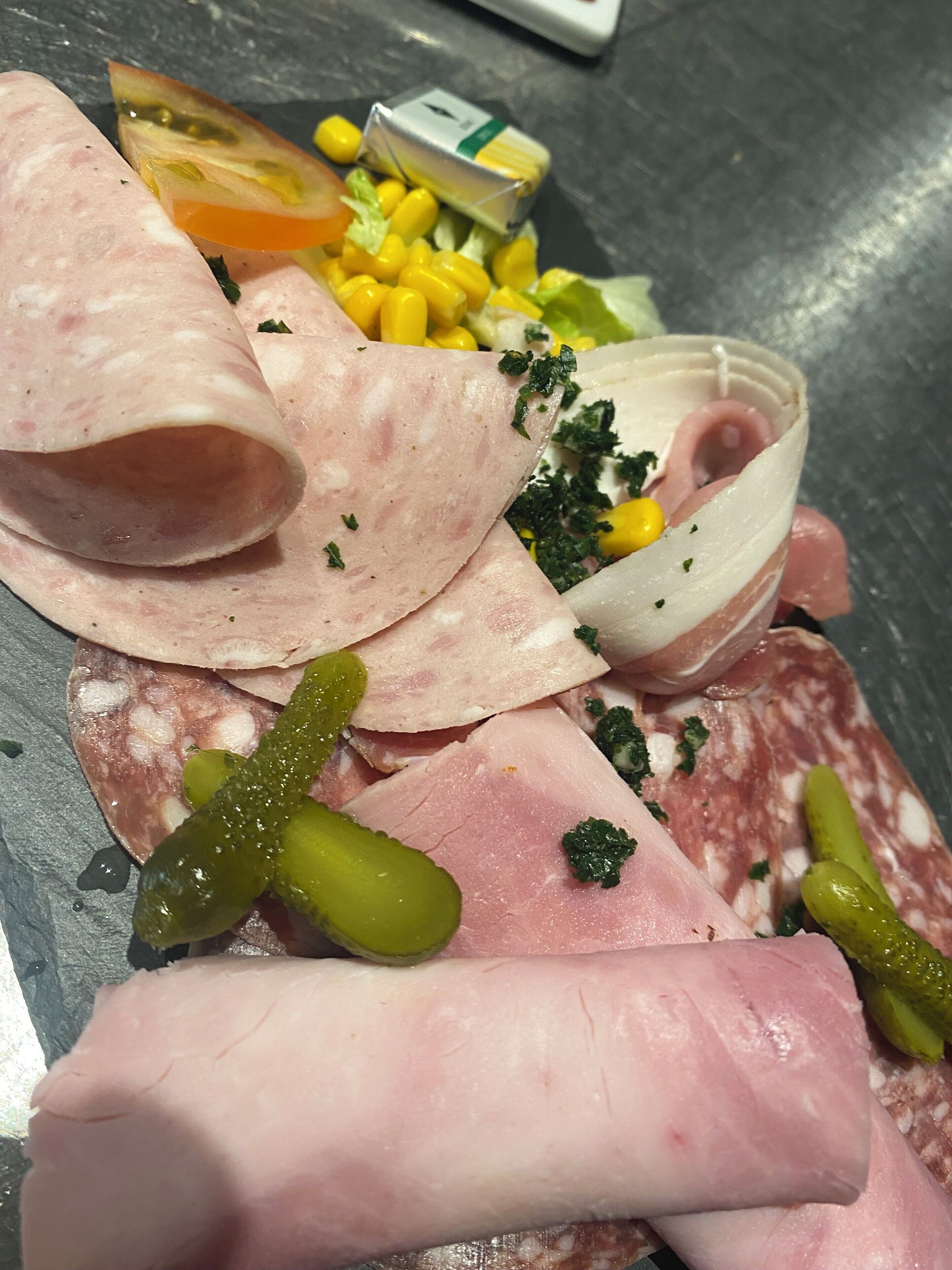 Diverse vleessoorten, zoals plak ham, salami en parmaham, vergezeld van augurken, mais en een kleine schaal groen.