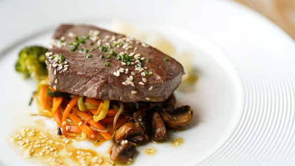 A plate with a steak topped with sesame seeds and chopped herbs, accompanied by sautéed mushrooms and carrots.