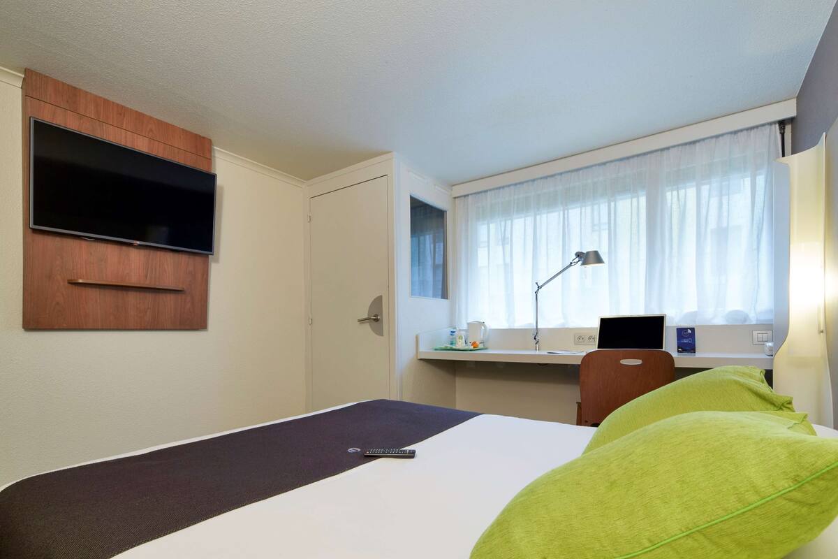Une chambre d’hôtel avec un lit, un bureau équipé d’un ordinateur, un téléviseur fixé au mur et une fenêtre avec des rideaux.