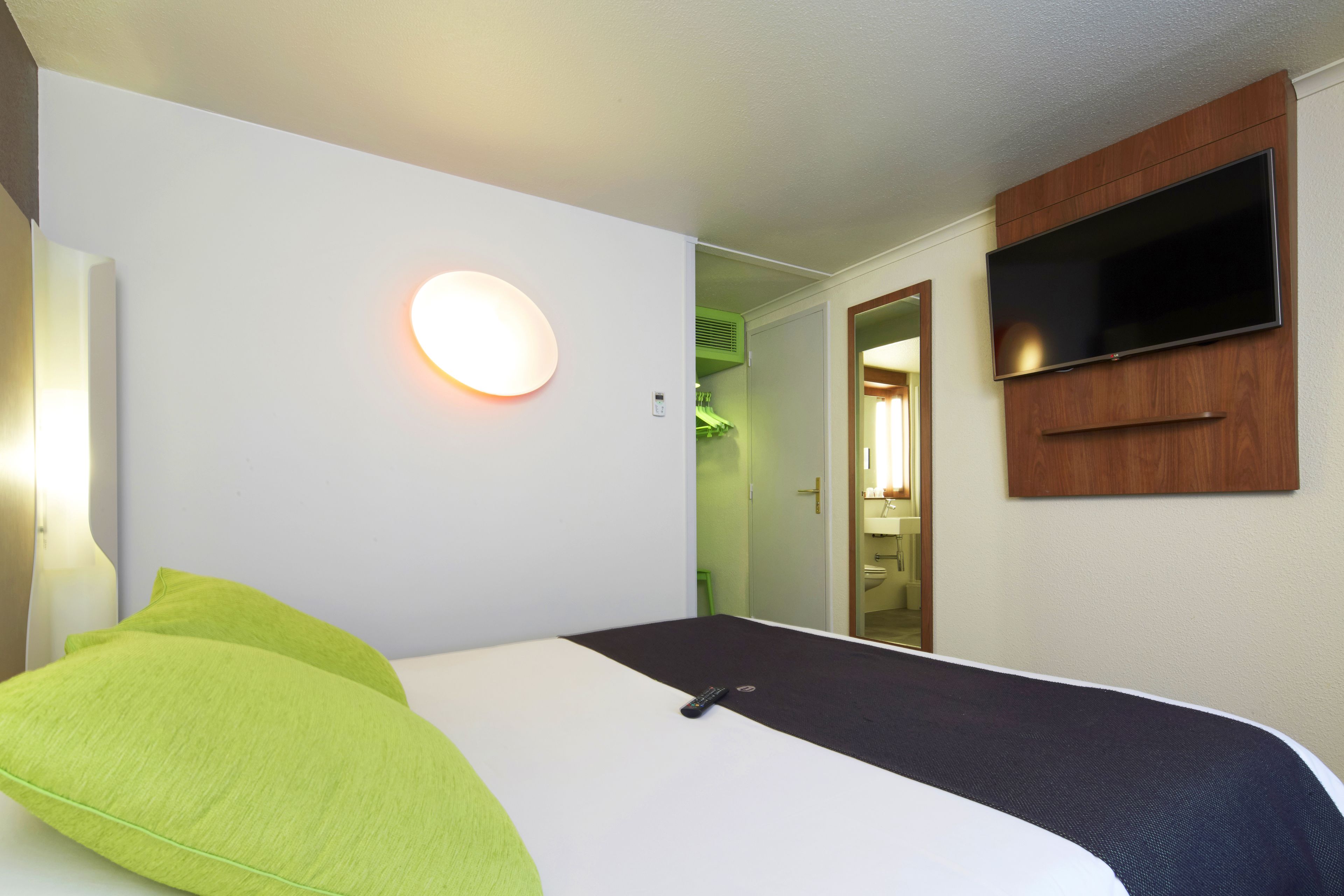 A hotel room with a bed, a flat-screen TV mounted on the wall, and a round wall light.