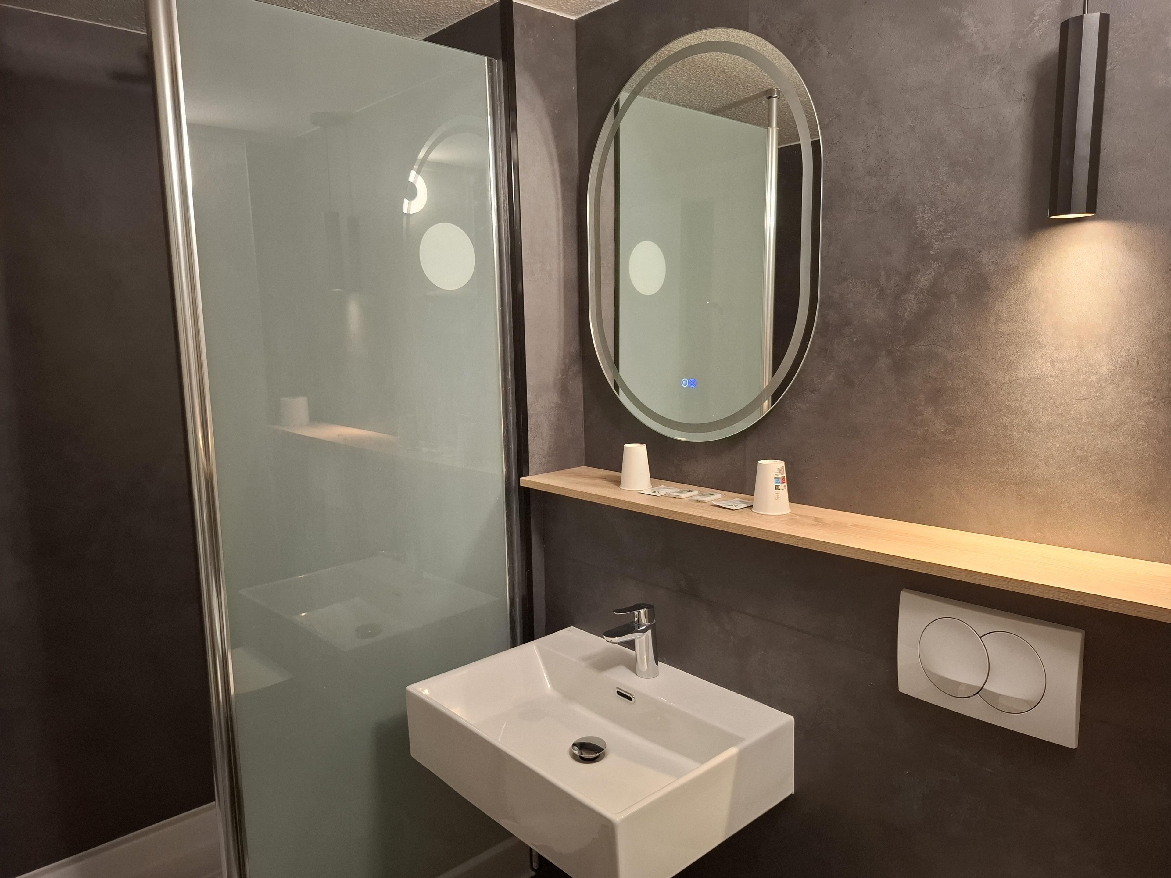 A modern bathroom with a glass shower enclosure, a white sink with a chrome faucet, and a mirror above a wooden shelf.