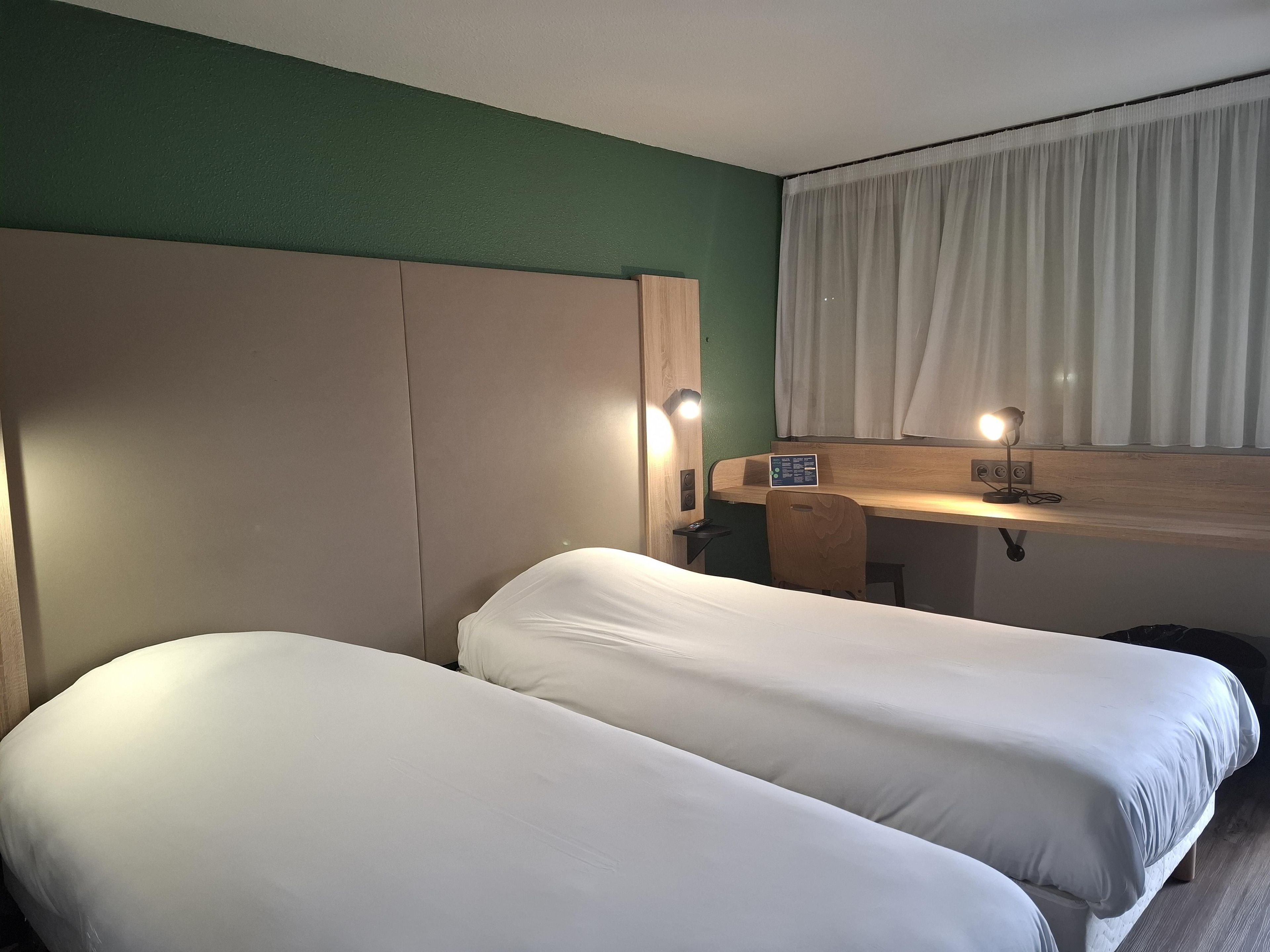 Une chambre d’hôtel avec deux lits, un bureau, une chaise, une fenêtre avec des rideaux et un luminaire fixé au mur.