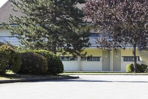 Un bâtiment jaune avec une allée d’accès fermée par un portail, entouré d’arbres et d’arbustes.