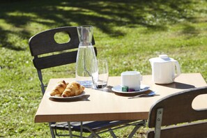Un montaje para picnic con una mesa, sillas, croissants, una botella de agua, un vaso, una taza, una tetera y una cuchara sobre un área cubierta de césped.