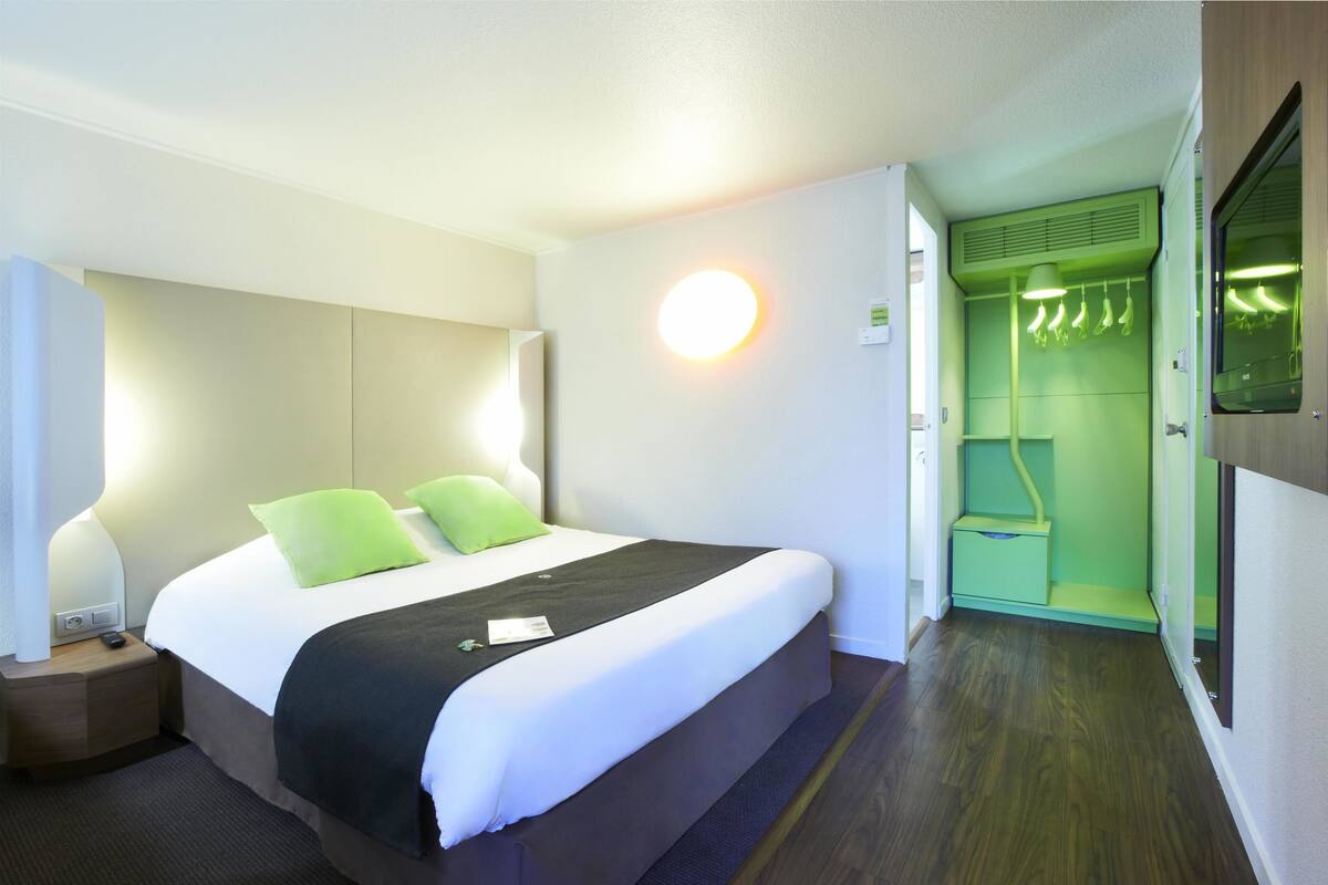 Une chambre d’hôtel avec un grand lit, une table de chevet, une lampe fixée au mur et une salle de bain aux tons verts.
