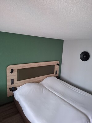 Una cama individual con cabecera de madera, una mesita de noche con un control remoto y un altavoz fijado a la pared.