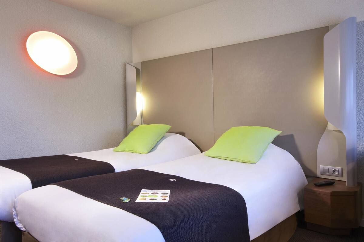 Une chambre d’hôtel avec deux lits, chacun étant équipé d’un oreiller et d’une couverture. Il y a une table de chevet avec une lampe et une télécommande.