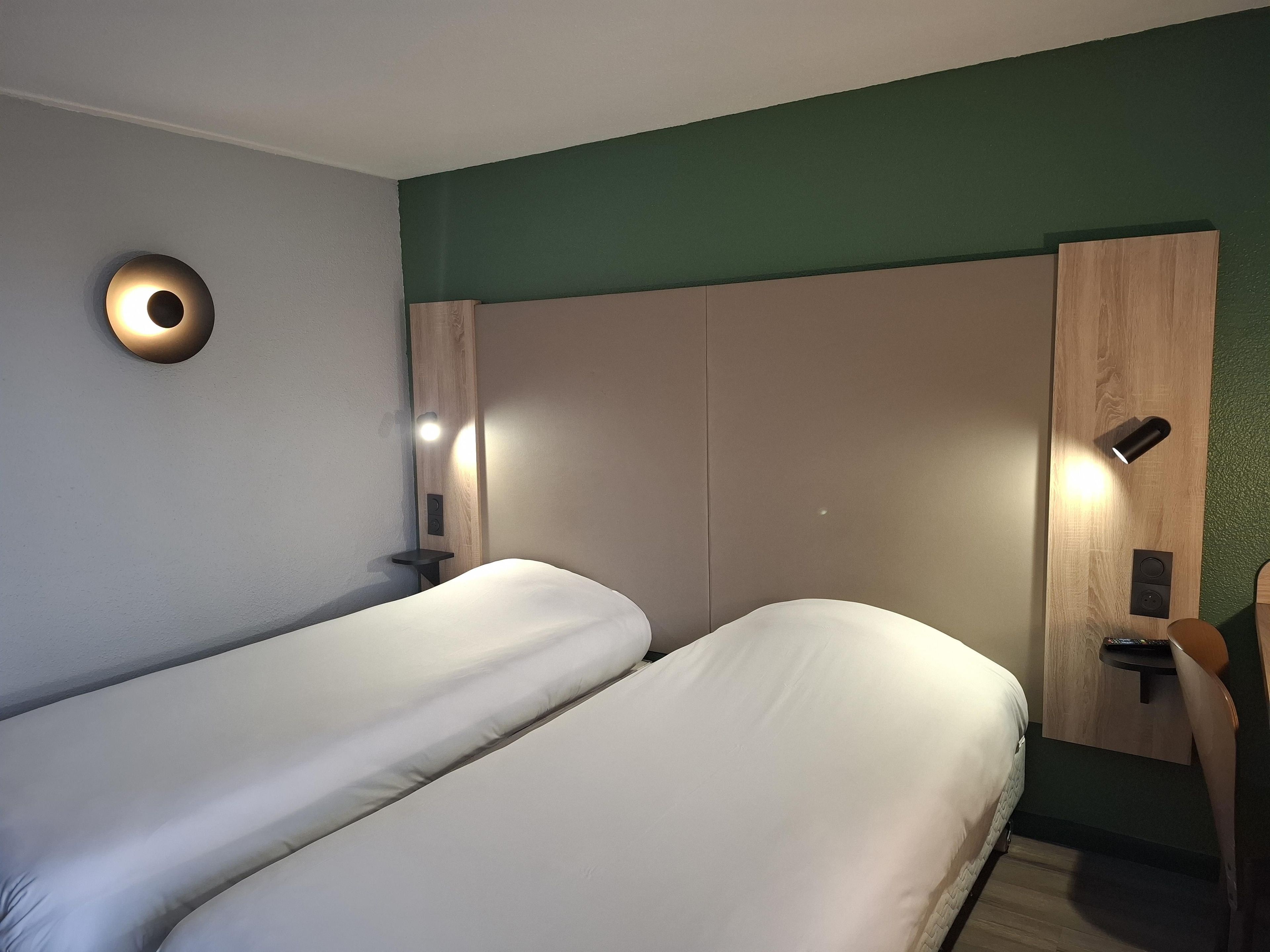 A hotel room with two single beds, a wooden wardrobe, a small bedside table, and a wall-mounted light fixture.