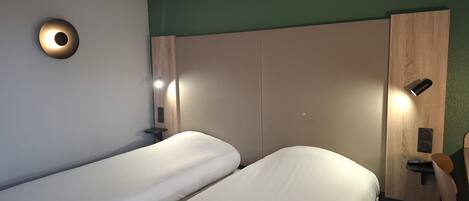A hotel room with two single beds, a wooden wardrobe, a small bedside table, and a wall-mounted light fixture.