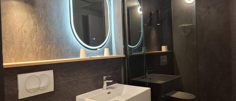 Un baño moderno con un lavamanos blanco, un espejo con iluminación perimetral y una zona de ducha con mampara de vidrio.