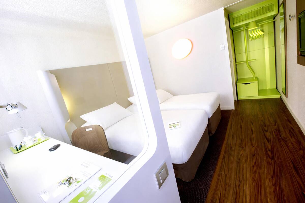 Quarto de hotel com duas camas, uma escrivaninha com luminária e corredor iluminado em tons de verde.