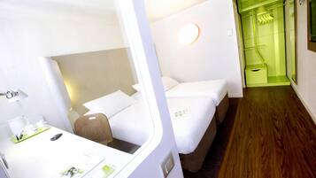 A hotel room with two beds, a desk with a lamp, and a green-lit hallway.