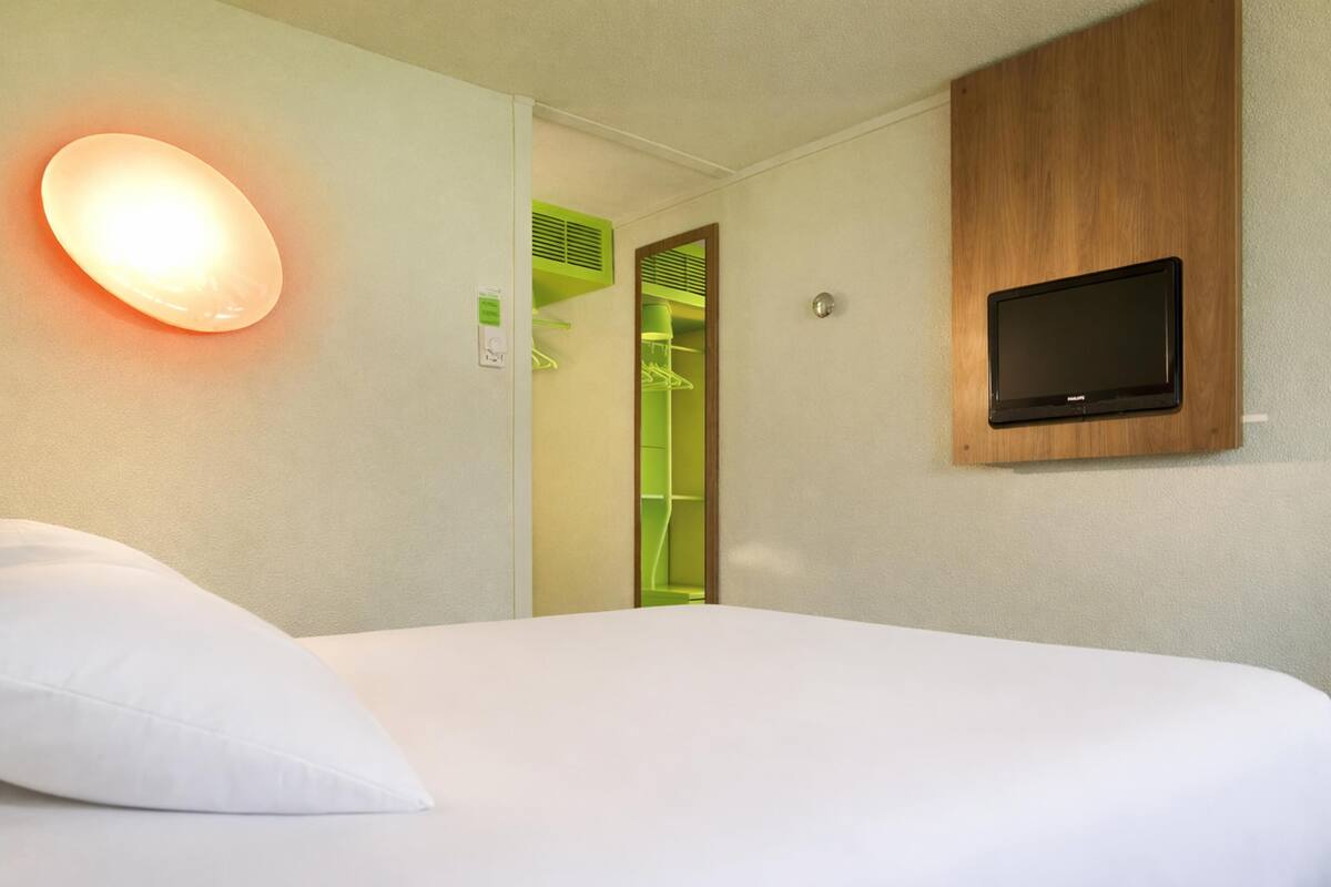 Quarto de hotel com cama, televisão fixada na parede, luminária e armário verde.