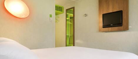 Una habitación de hotel con una cama, un televisor montado en la pared, una lámpara y un armario verde.