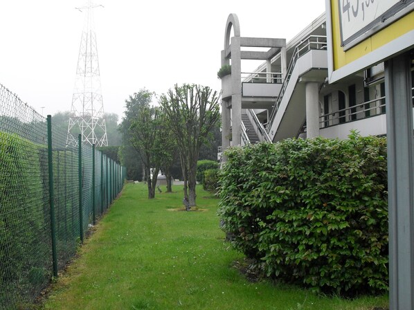 A pathway flanked by a green fence and a hedge, leading to a modern building with a staircase and a yellow sign. - Premiere Classe Hotel Liege (Liege)