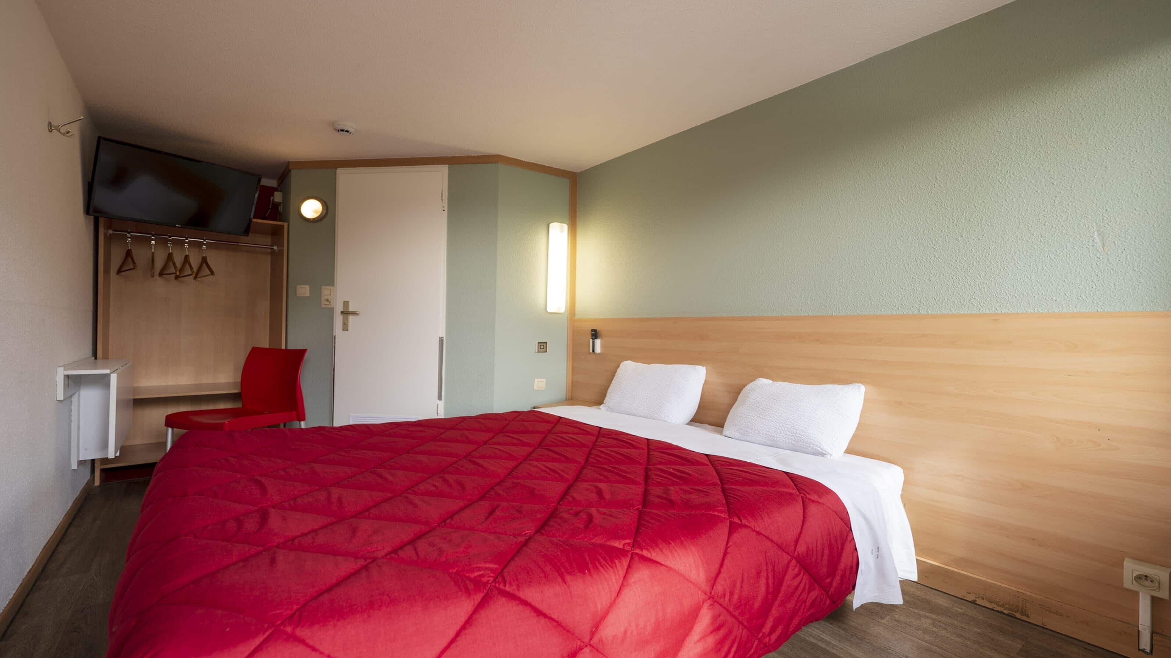 Habitación de hotel con una cama grande, una silla roja, un armario y un televisor en la pared.