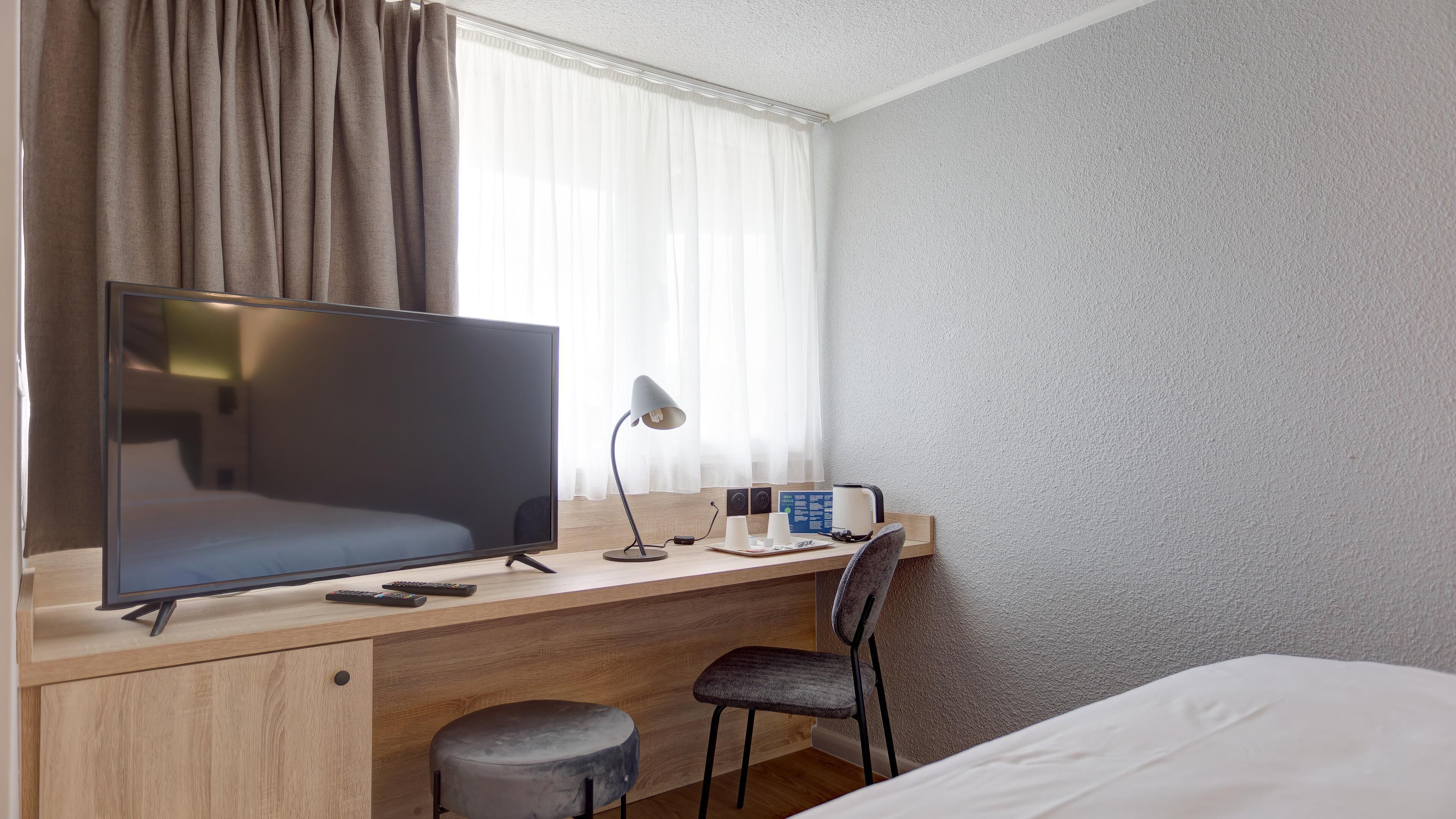 A hotel room with a bed, a desk with a television, a chair, a lamp, and a window with curtains.