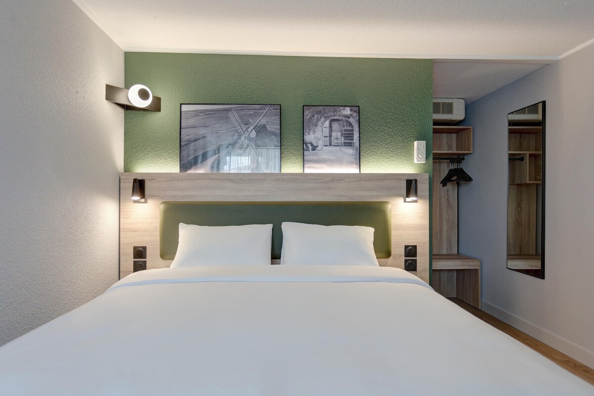 Habitación de hotel con una cama, dos cuadros colgados en la pared, una mesita de noche y un espejo.